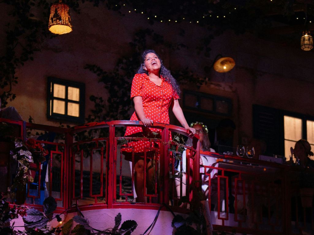 빨간 물방울 무늬 드레스를 입은 여성이 발코니에 서서 창문과 덩굴이 희미하게 빛나는 배경으로 노래를 부르고 있습니다.