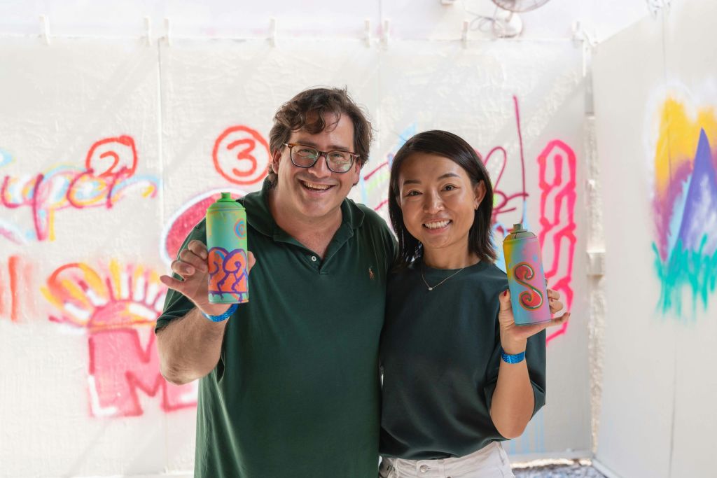 화려한 색상의 스프레이 페인트 캔을 들고 활기찬 그래피티 아트가 있는 벽 앞에 서 있는 두 사람이 미소를 짓고 있습니다.