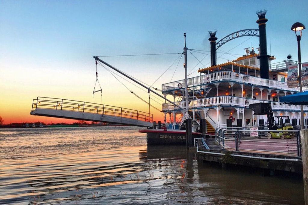 "크리올 퀸" 이라는 이름의 도킹 패들휠 스팀보트로, 석양을 배경으로 왼쪽에는 높이 솟은 보딩 램프가 있습니다.