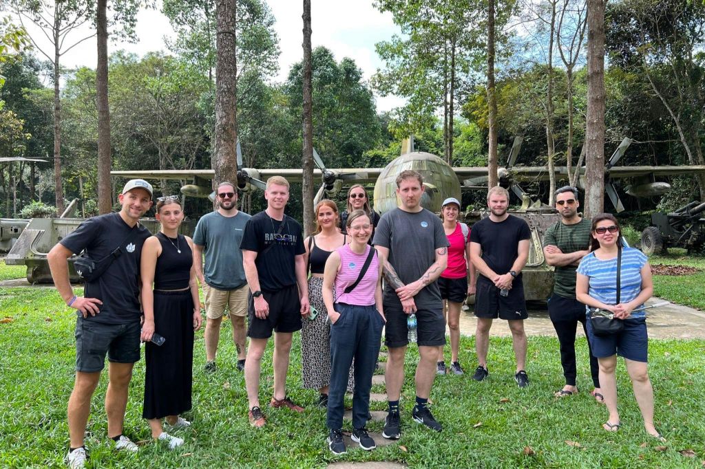한 무리의 사람들이 나무와 오래된 군용기를 배경으로 잔디밭에 서 있습니다.