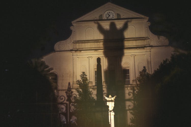 팔을 쭉 뻗은 불이 켜진 조각상은 밤에 교회 정면에 커다란 그림자를 드리우며, 나무와 시계가 보입니다.