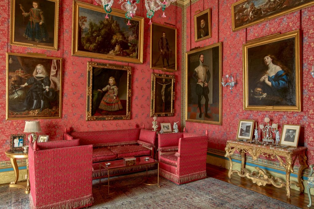 빨간색 벽지, 커다란 액자 그림, 빨간색 소파, 액자 사진을 보여주는 금색 콘솔 테이블이 있는 화려한 객실입니다.