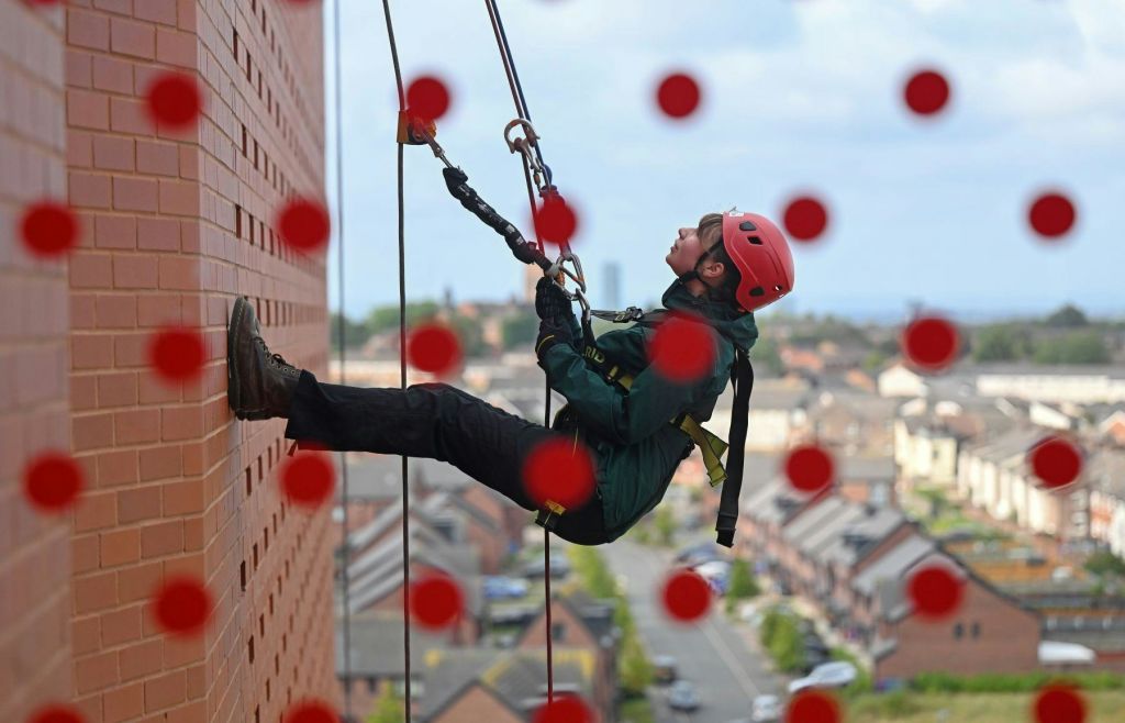 빨간 헬멧과 하네스가 달린 벽돌 벽을 따라 내려가는 안전 장비를 착용한 사람. 빨간색 점이 이미지를 덮습니다. 아래 주택이 보입니다.