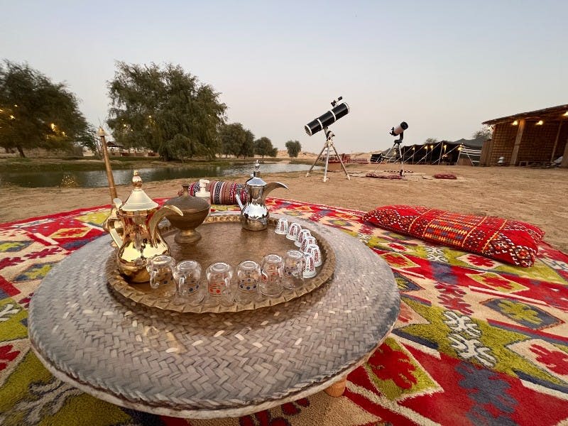 직조된 트레이 위에 찻주전자와 안경을 놓은 낮은 테이블, 화려한 카펫, 하늘을 가리키는 망원경이 있는 야외 공간.