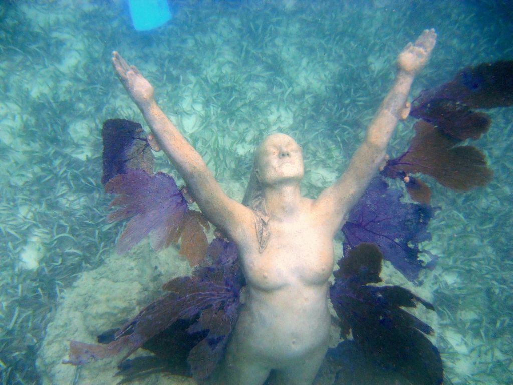 바다 바닥에 해초와 같은 요소로 둘러싸인 두 팔을 들어 올린 인간 형상의 수중 동상.