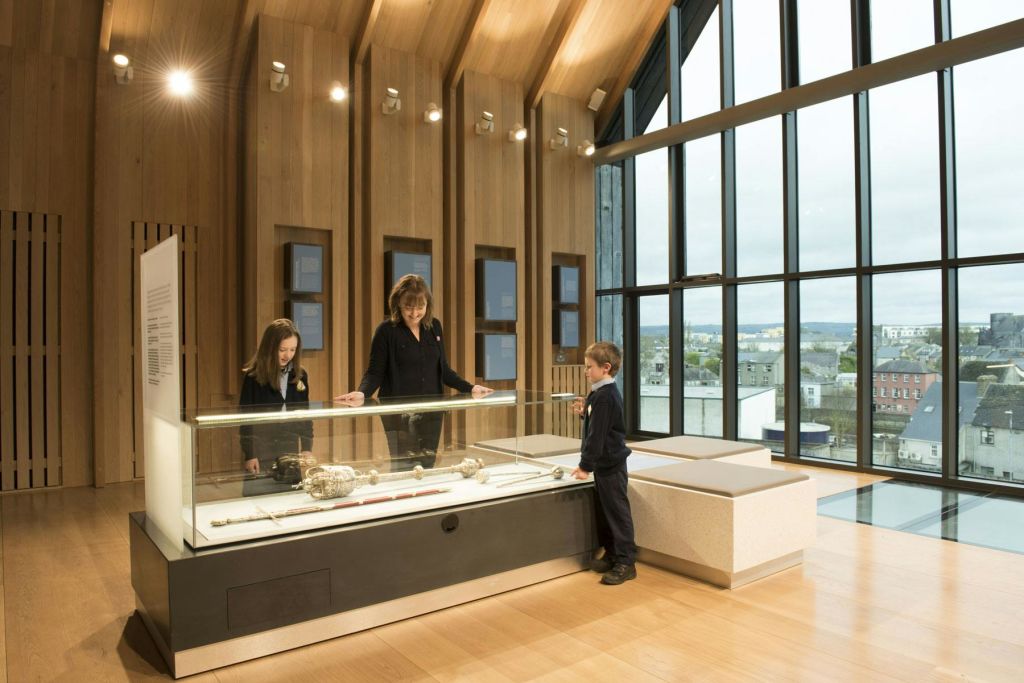 커다란 창문이 있는 현대적인 목조 패널 객실 내부에서 박물관 진열 케이스의 유물을 살펴보는 세 사람.
