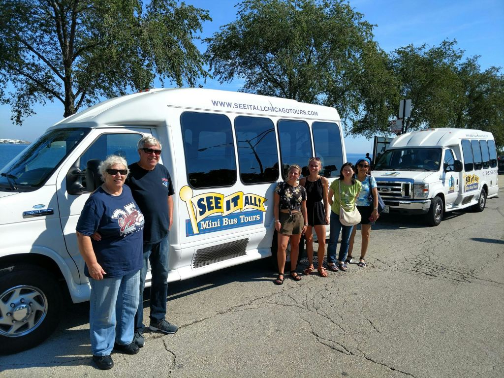 나무가 우거진 호숫가 근처에서 'See It All 미니 버스 투어' 라고 적힌 흰색 투어 밴 앞에서 한 무리의 사람들이 포즈를 취하고 있습니다.