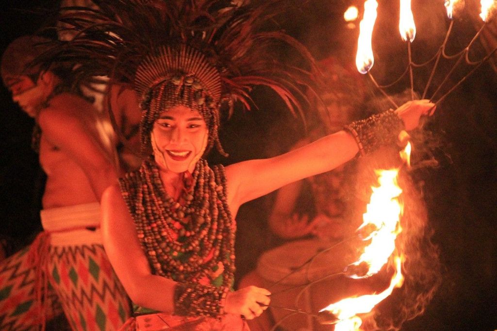 머리 장식이 달린 전통 복장을 한 사람이 횃불을 들고 웃으며 춤을 추고 있습니다. 비슷한 의복을 입은 다른 사람들이 배경에 있습니다.