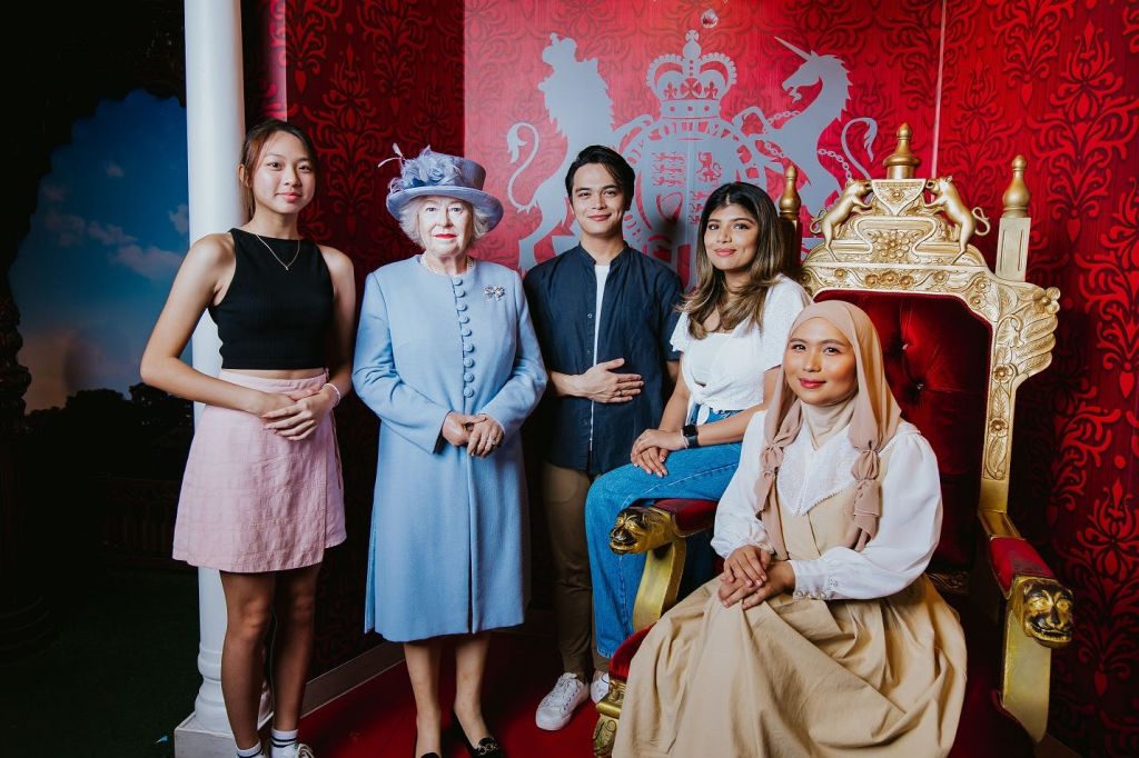 파란색 의상과 모자를 쓰고 왕실 문장이 있는 빨간색 배경에 세팅된 여성의 왁스 피규어를 들고 네 명이 포즈를 취하고 있습니다.