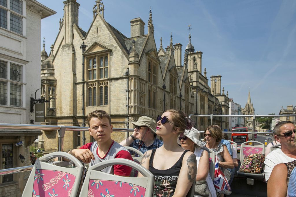역사적인 석조 건물과 맑은 하늘을 배경으로 투어 버스의 꼭대기 갑판에 앉아 있는 사람들.