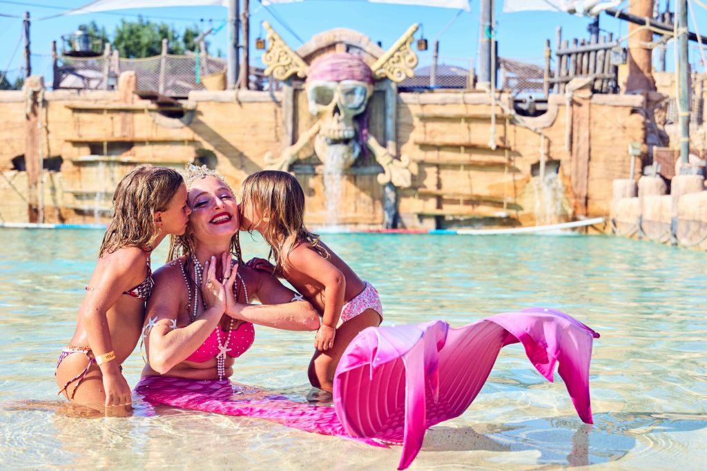 분홍색 인어공주 의상을 입은 한 여성이 해적 장식이 있는 워터파크에서 두 아이가 뺨에 키스하고 얕은 물에 앉아 있습니다.