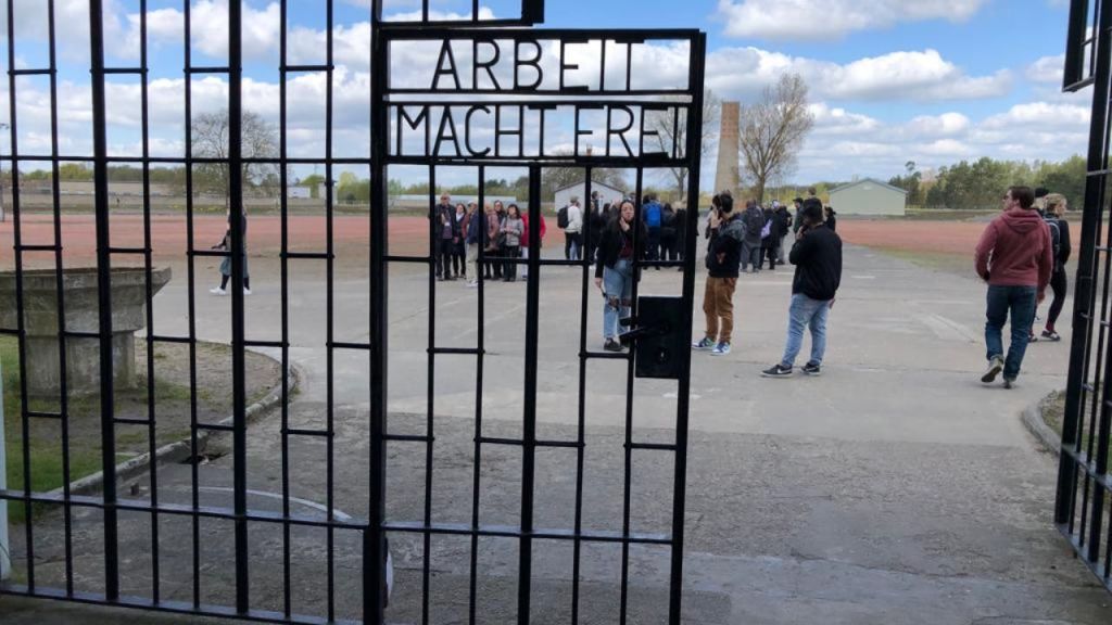 한 무리의 사람들이 "ARBEIT MACHT FREI" 라고 새겨진 철문 뒤에 서 있습니다. 하늘이 부분적으로 흐리다.