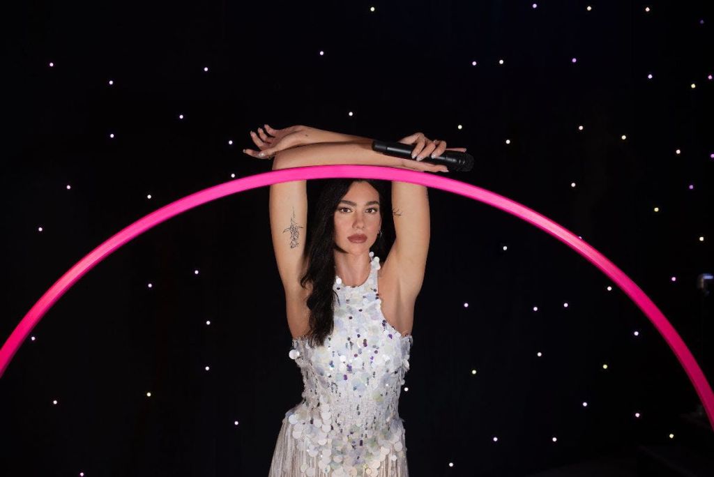 시퀸 드레스를 입은 여성이 분홍색 훌라 후프가 박힌 마이크를 머리 위로 들고 있습니다. 별이 빛나는 배경.