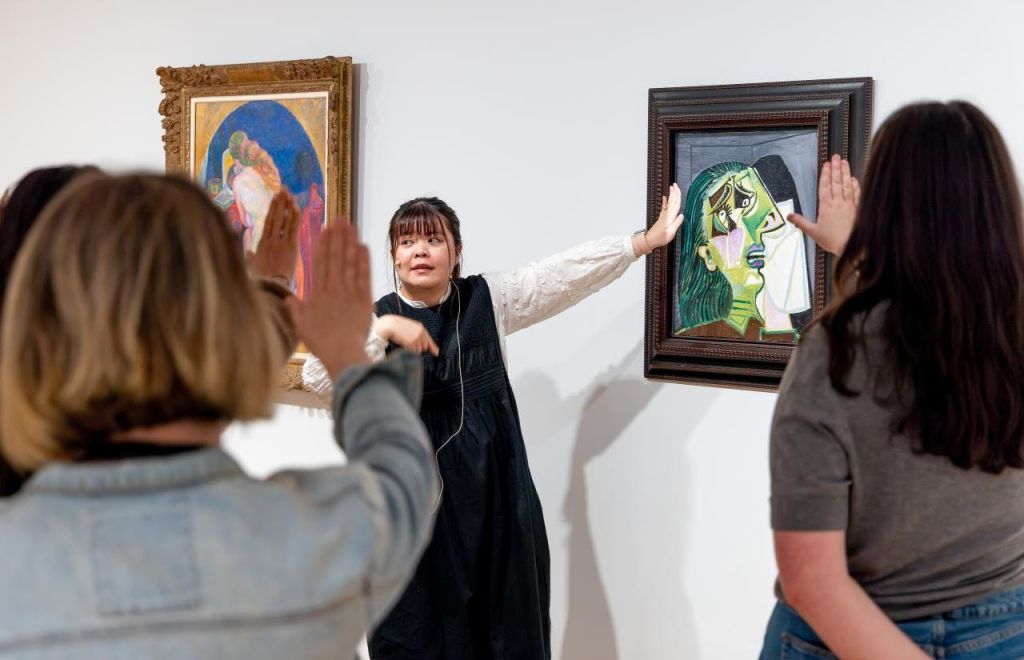 두 개의 액자로 된 예술품 사이에 서서 몸짓을 하는 사람으로, 전경에 있는 다른 사람이 손을 들고 있습니다.