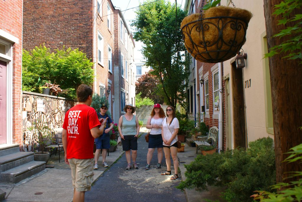 벽돌 건물과 녹지가 늘어선 골목에서 투어 가이드가 네 명과 이야기를 나눕니다.