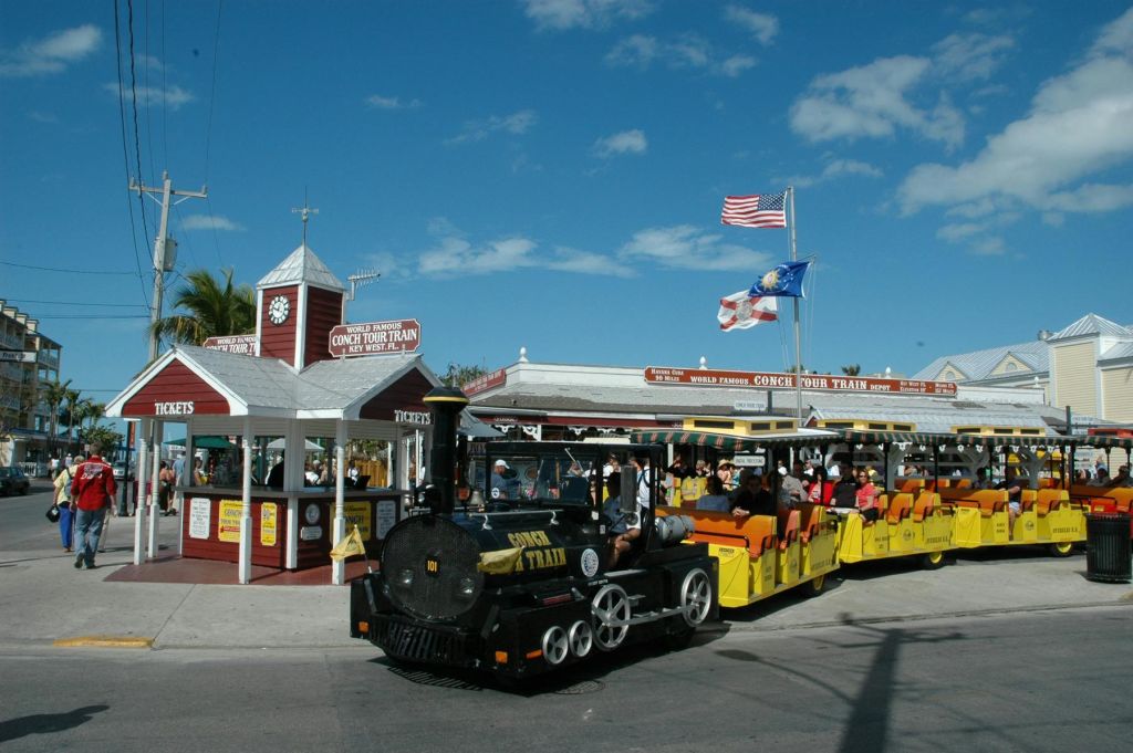 노란색 관광열차가 소라 투어 트레인 (Conch Tour Train) 이라고 적힌 빨간색과 흰색 부스 앞에 승객을 태우고 있습니다. 깃발은 푸른 하늘을 향해 머리 위로 날아갑니다.