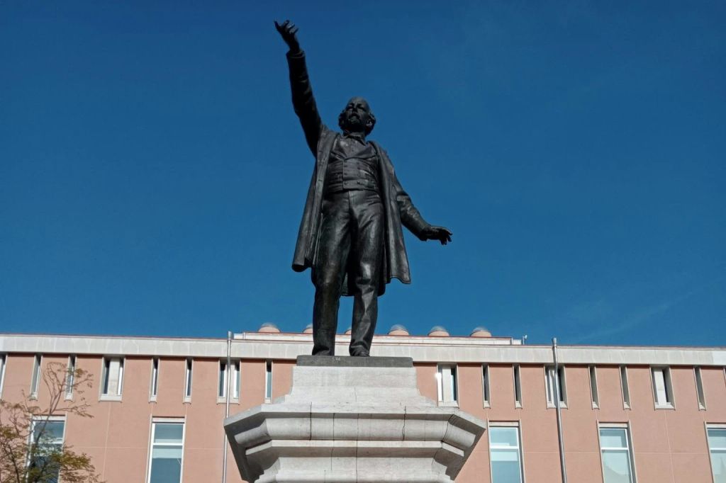 맑고 푸른 하늘을 배경으로 현대적인 건물 앞의 돌 받침대 위에 팔을 들어 올린 남자의 청동 동상.