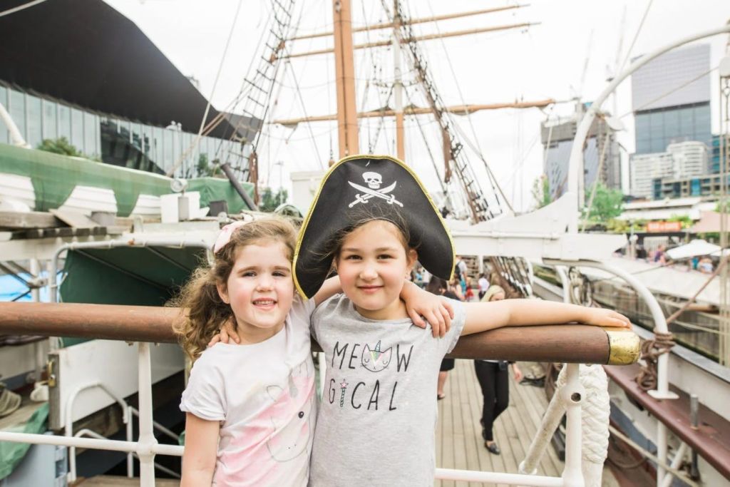 해적 모자를 쓴 두 어린이가 배 갑판에 서 있습니다. 배경에는 역사적인 선박과 리깅이 보입니다.