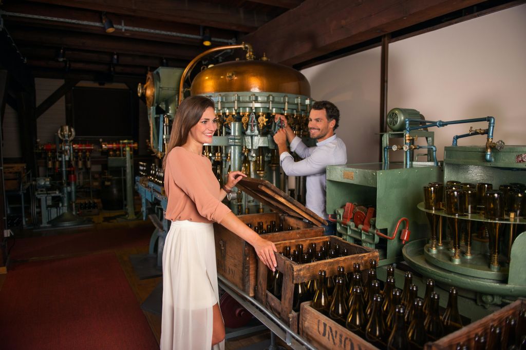 한 남녀가 산업용 병입 기계에서 맥주를 병에 담고 웃으며 빈 병으로 채워진 나무 상자를 다루고 있습니다.