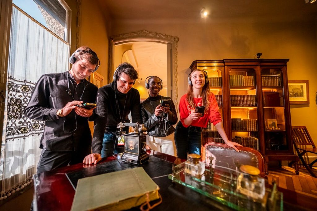 헤드폰을 착용한 세 사람이 나무 캐비닛과 책상 위에 놓인 앤티크한 물건이 놓인 역사적인 방을 탐험합니다.