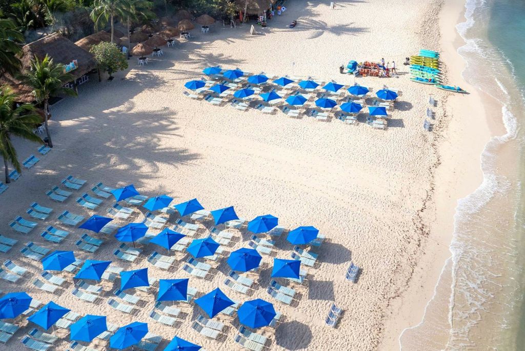 수많은 파란 우산과 일광욕 의자가 줄지어 있는 해변의 공중 전망. 배경에는 나무와 오두막이 있습니다.