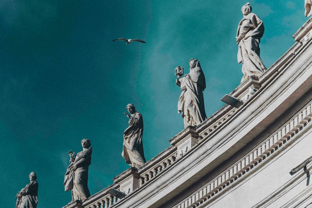 청록색 하늘 배경의 곡선형 건물 꼭대기에 있는 동상. 조각상 근처에서 새가 하늘을 날고 있습니다.