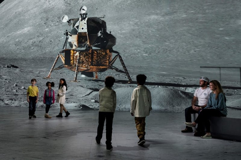 두 명의 어린이가 근처에 서 있는 동안 두 명의 성인이 벤치에 앉아 박물관의 대형 달 모듈 디스플레이를 보고 있습니다.