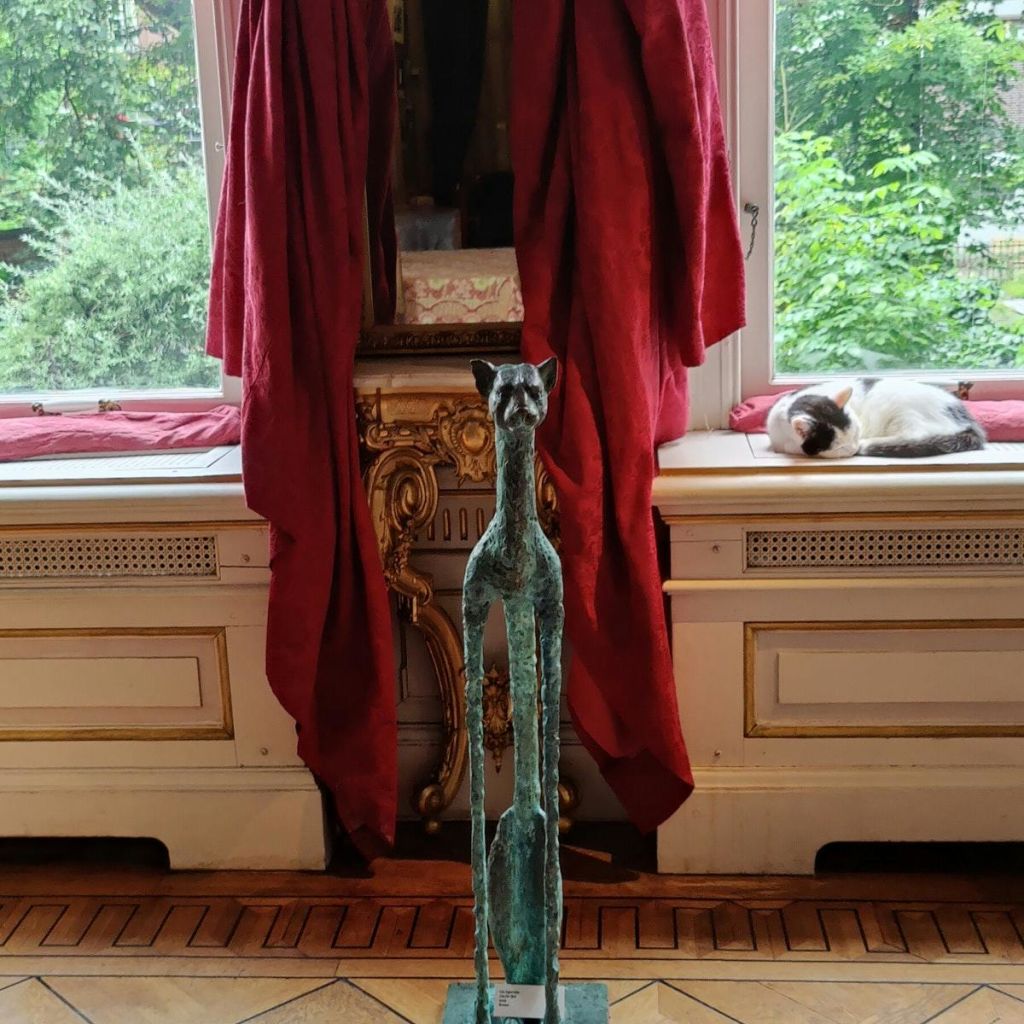 커다랗고 가느다란 고양이 조각상이 커다란 창문 앞에 서 있습니다. 창문턱에서 고양이가 낮잠을 자고 있습니다.