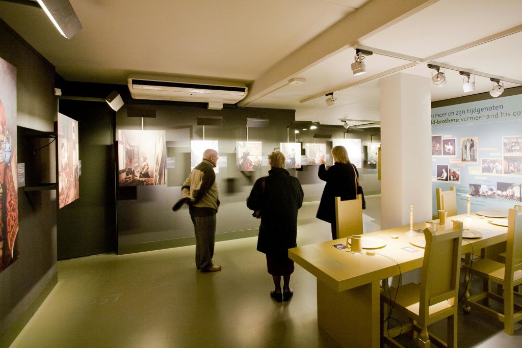 조명이 희미한 갤러리에서 액자에 담긴 예술작품을 감상하는 세 사람. 개체가 있는 테이블은 오른쪽 앞에 있습니다.