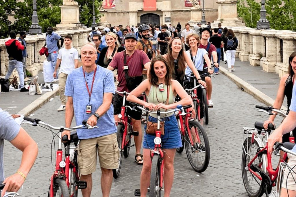빨간색 자전거를 탄 사람들이 인파와 역사적인 건축물을 배경으로 자갈길에서 미소를 짓고 포즈를 취하고 있습니다.