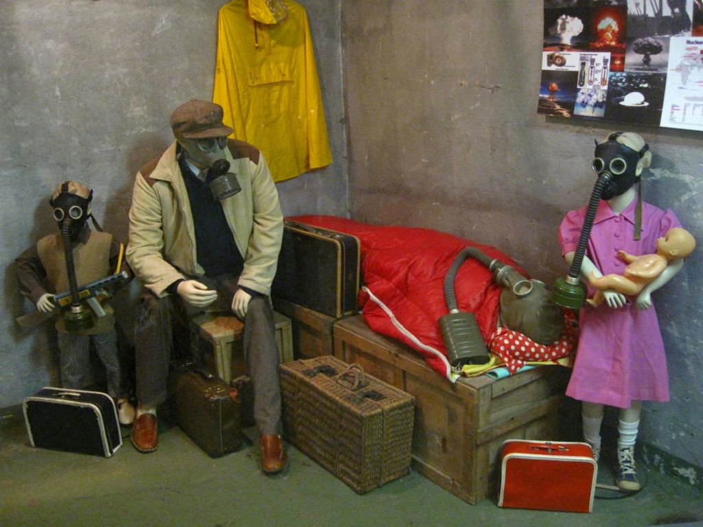 방독면을 쓴 마네킹 두 개가 여행 가방이 놓인 방에 앉아 있고, 벽에 노란색 레인코트가 걸려 있습니다. 나무 상자에 빨간색 침낭이 있습니다.