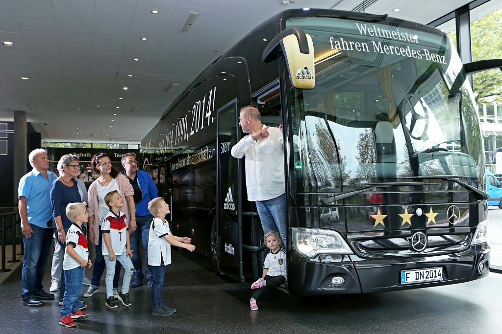 건물 안에는 어른과 어린이 무리가 검은색 버스를 타고 서 있습니다. 일부 어린이는 검은색과 빨간색으로 포인트를 준 흰색 셔츠를 입고 있습니다.