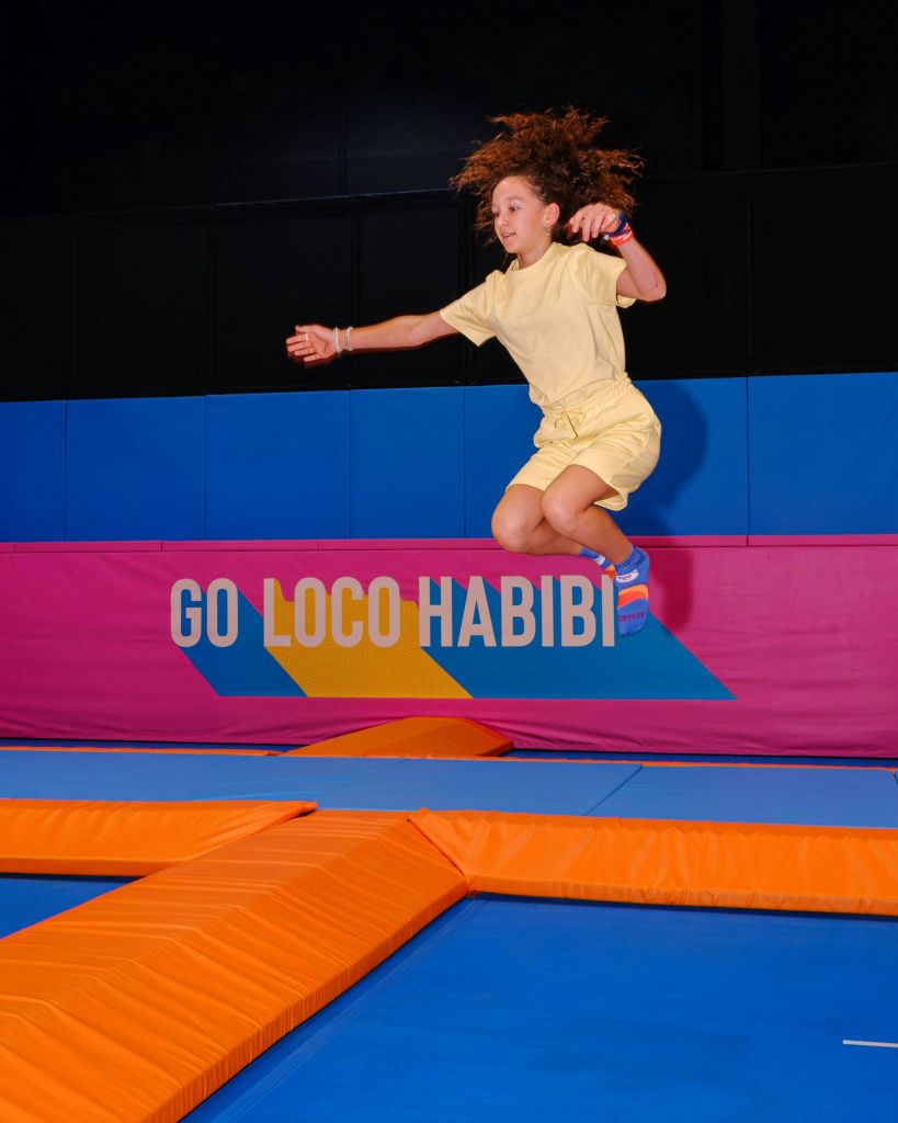 한 젊은이가 노란색 의상을 입고 실내 트램펄린에서 뛰어내리고 있습니다. 배경에는 "GO LOCO HABIBI" 라고 쓰인 분홍색 배너가 있습니다.