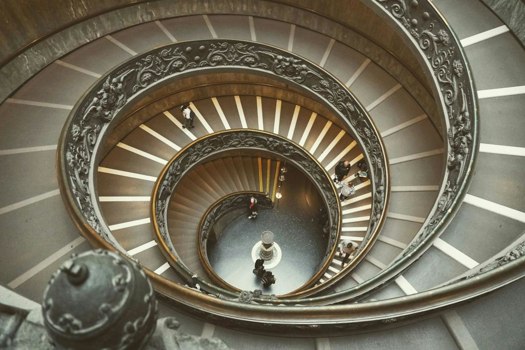 위에서 바라본 화려한 난간이 있는 나선형 계단. 사람들은 내려오고 올라갑니다. 중앙에는 작은 조각상이 있습니다.