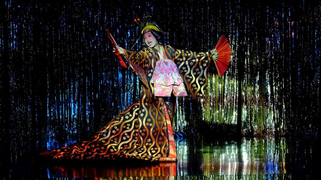 반짝이는 파란색 무대에서 빨간색 팬을 들고 팔을 쭉 뻗은 전통 의상과 메이크업을 입은 연주자.