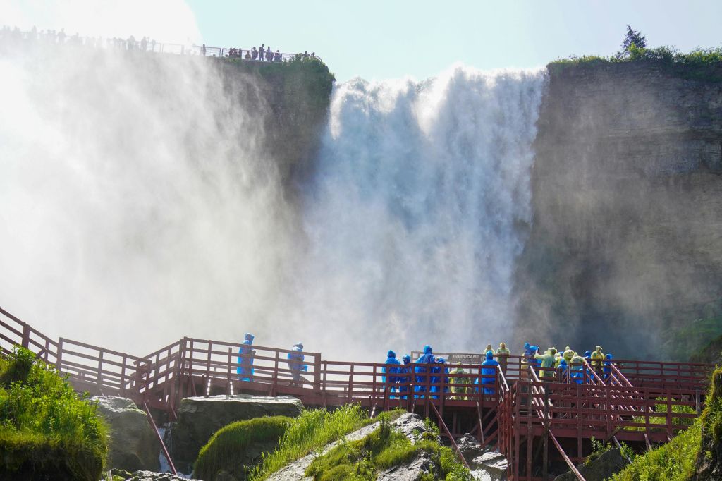 파란색과 노란색 비옷을 입은 사람들이 커다란 폭포 근처의 빨간색 보드워크를 걷고 있으며, 주변에는 안개가 솟아 있습니다.