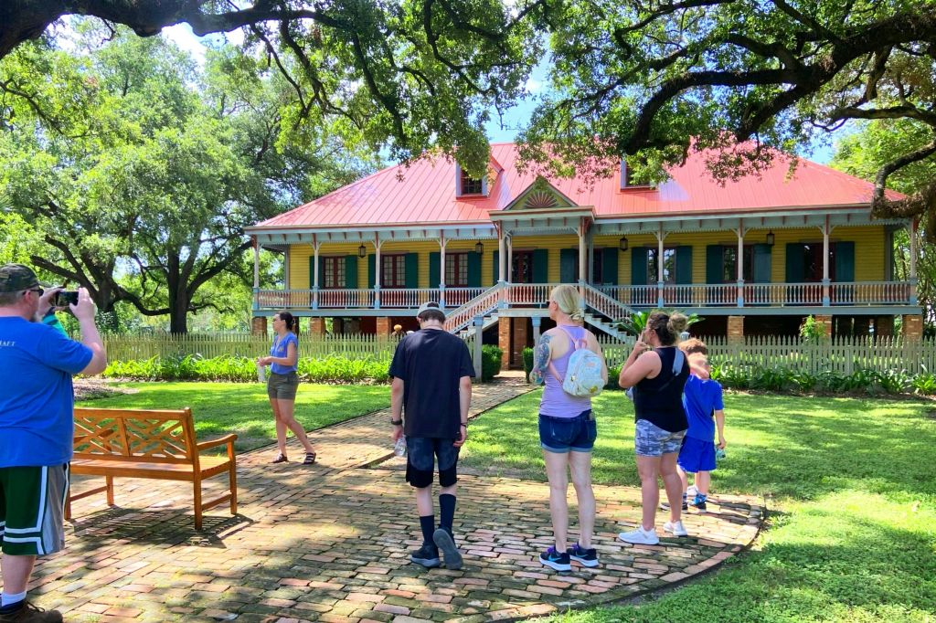 한 무리의 사람들이 나무로 둘러싸인 빨간색 지붕과 포장된 현관이 있는 크고 화려한 집 앞 통로 근처에 서 있습니다.