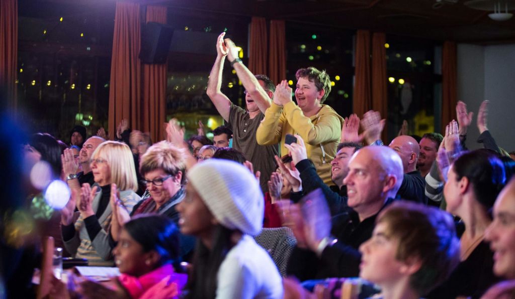 한 무리의 사람들이 실내에서 박수를 치며 환호하고 있으며, 두 사람이 서서 흥분한 듯 손을 들고 있습니다.