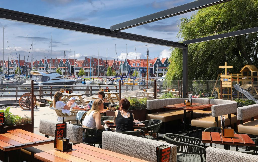 정박지 옆 레스토랑의 야외 좌석 공간에는 사람들이 식사를 하고 화려한 집들이 배경으로 자리하고 있습니다.