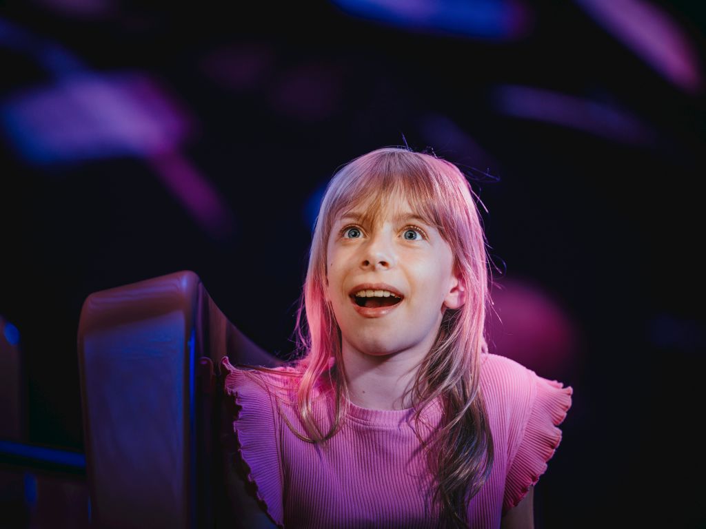 금발에 핑크색 상의를 입은 어린 소녀가 화려하고 흐릿한 조명을 배경으로 놀라움과 설렘을 선사합니다.