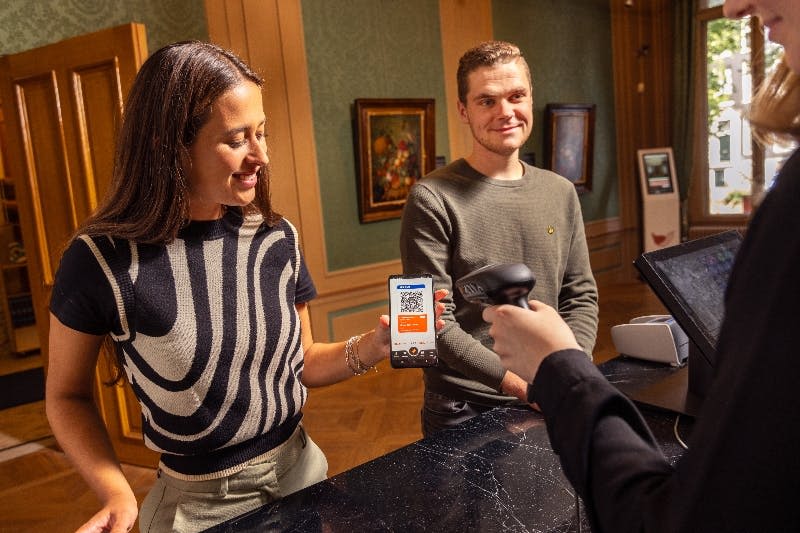 한 여성이 휴대전화로 스캔할 QR 코드를 제시하고, 한 남성이 옆에 서서 따뜻하게 빛나는 방에서 미소를 짓고 있습니다.