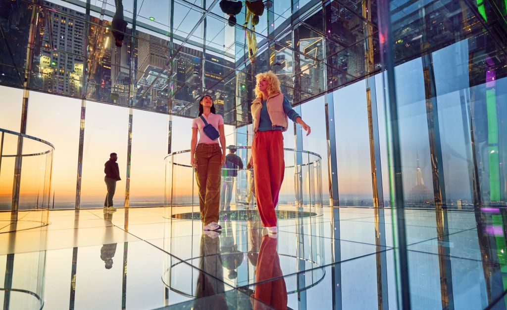 도시 전망과 석양의 다채로운 색채가 어우러진 거울의 방에 두 명의 여성이 서 있습니다. 한 남자가 배경을 관찰하고 있다.