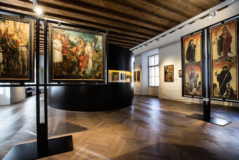 벽과 칸막이, 나무 바닥, 자연광이 들어오는 창문에 커다란 그림이 전시된 미술관.