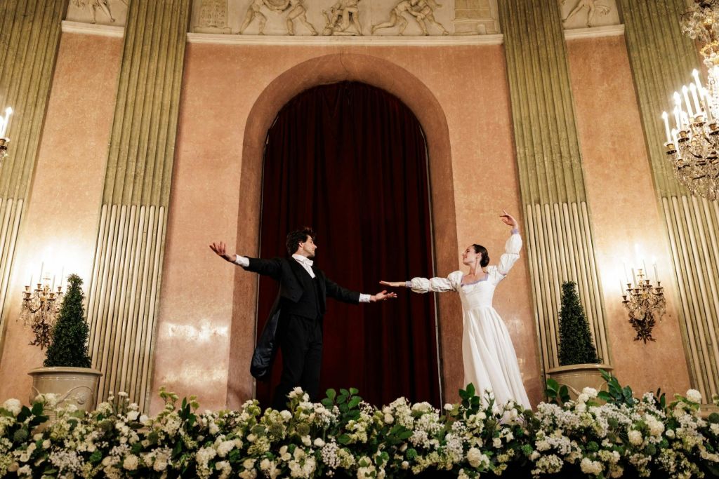 우아한 복장을 한 두 사람이 무대에서 꽃 장식과 붉은 커튼을 배경으로 춤을 추고 있습니다.