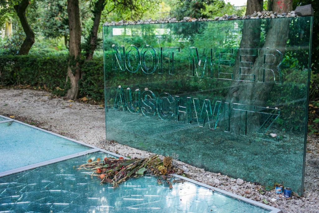 "Nooit Meer Auschwitz" 라는 글씨가 새겨진 반사 유리 기념관과 시든 꽃이 바닥에 놓여 있습니다. 나무는 배경에 있습니다.