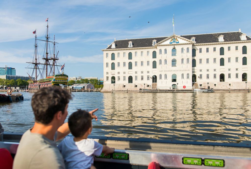 보트를 탄 어른과 어린이가 물 건너편에 있는 "Het Scheepvaartmuseum" 이라는 크고 유서 깊은 건물을 바라보고 있습니다.