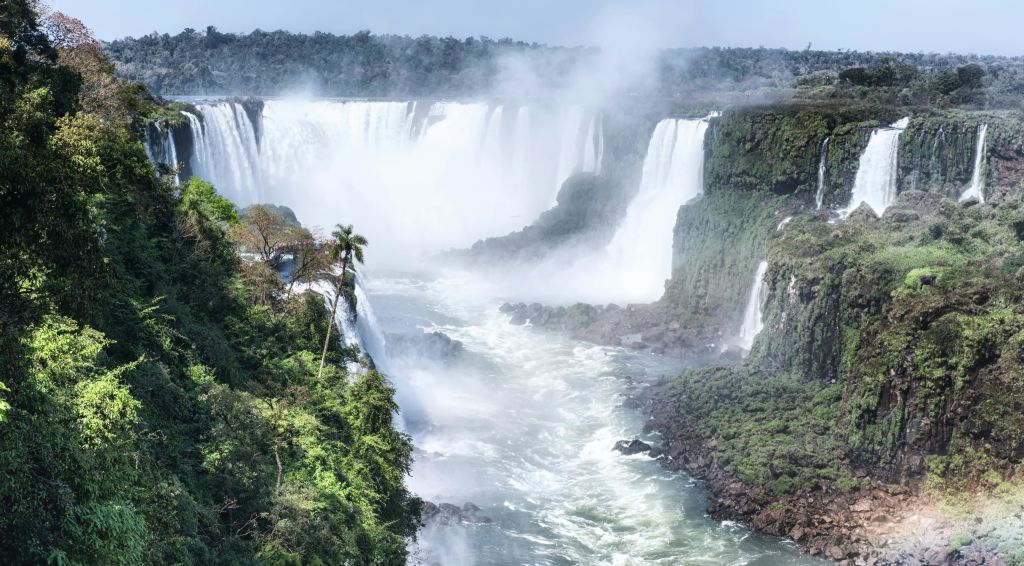 폭포는 울창한 녹음으로 둘러싸인 절벽 아래로 이어지며, 아래 강에서 안개가 솟아오릅니다. 빽빽한 숲이 배경으로 보입니다.