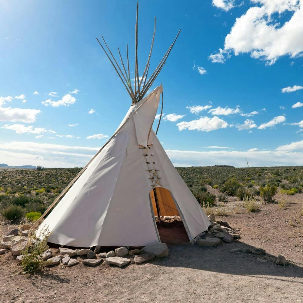 구름이 흩뿌려진 밝고 푸른 하늘 아래 사막 풍경에 하얀 티피가 서 있습니다. 돌들이 기지를 둘러싸고 있다.
