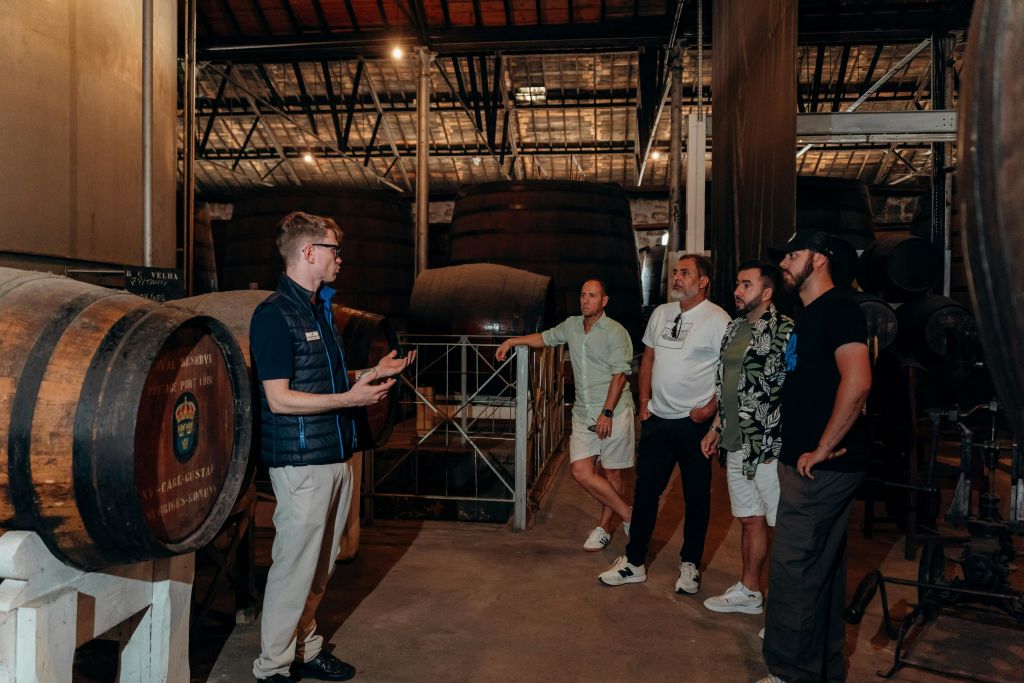 캐주얼한 복장을 한 5명의 남성이 커다란 나무 통으로 둘러싸인 증류소나 창고에 서서 대화를 나누고 있습니다.