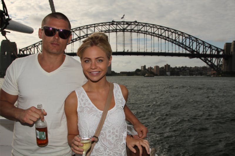 커다란 다리 앞에서 두 사람이 배를 타고 한 사람은 맥주를 들고 다른 한 사람은 샴페인을 들고 있습니다.